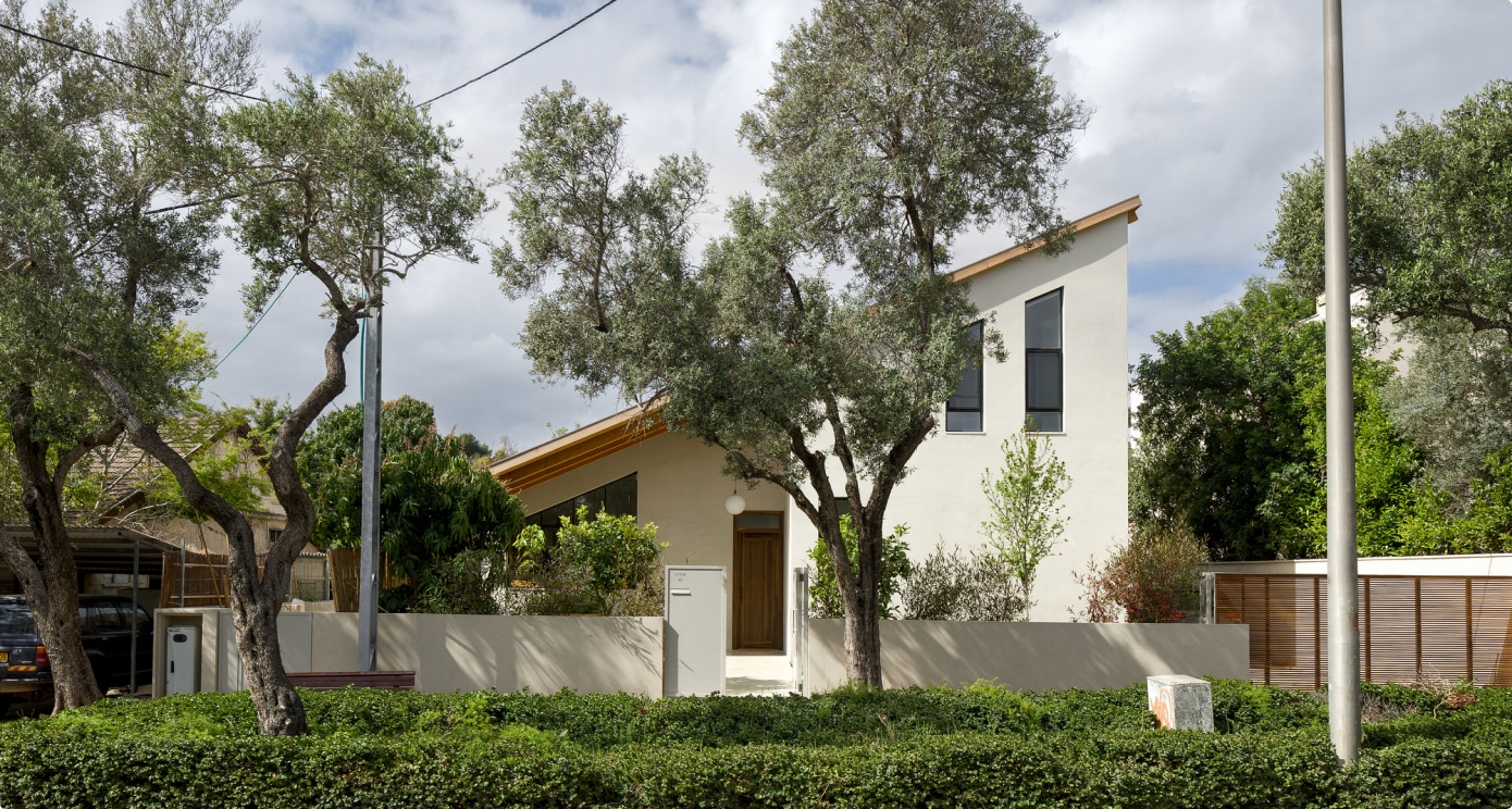Une maison blanche moderne au toit en pente se dresse derrière un muret, entourée d'arbres et de verdure, sous un ciel partiellement nuageux. On aperçoit de hautes fenêtres étroites sur la façade avant.