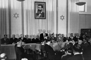 A group of men sit at a long table in a formal room with two Star of David symbols and a large portrait above. One man stands and speaks to an audience.