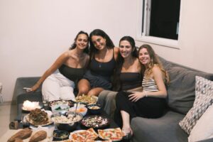 Four women sit on a gray couch, smiling at the camera with a coffee table in front of them filled with various dishes, including pizza, salads, bread, and snacks. The atmosphere is casual and friendly.