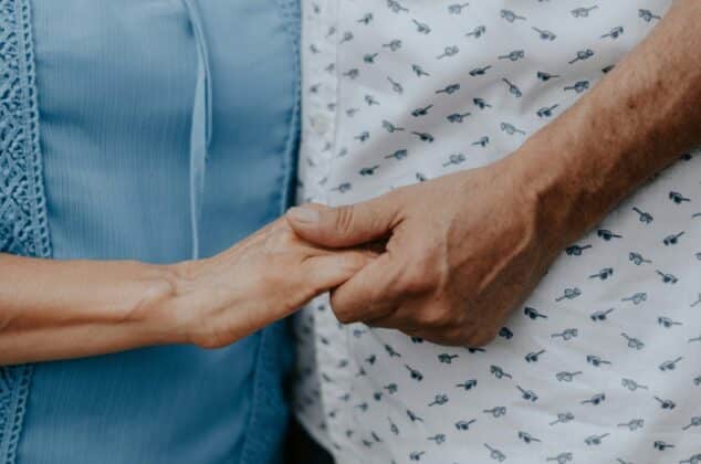 Two people stand close together, gently holding hands. One wears a light blue top, and the other wears a white shirt with a small black pattern. The image conveys a sense of warmth and connection.