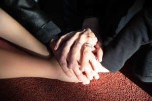 Three hands, some wearing rings, are stacked together in sunlight on a reddish textured surface, suggesting unity or support.