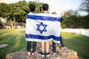Three children stand outdoors on a rock, their backs facing the camera, holding an Israeli flag draped behind them. Green trees and buildings are visible in the background.