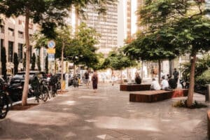 People sit and walk along a sunny city plaza with trees, benches, and bicycles. Tall buildings and shops line the background.